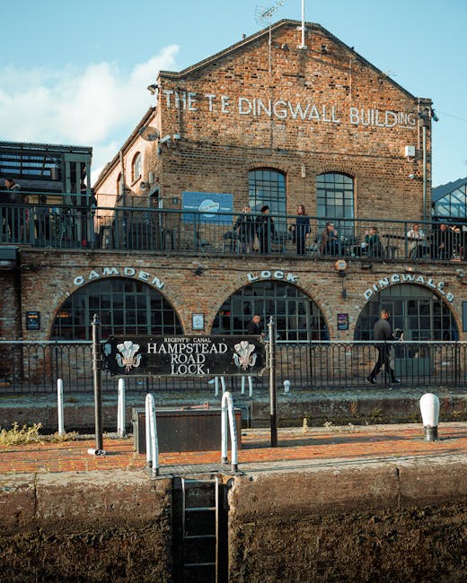 Exterior view of the historic Dingwall Building, a brick structure with large arched windows on the ground floor and rectangular windows on the upper floor, located alongside Regent's Canal in Camden. The upper balcony features a black metal railing, with several people standing and engaging in conversations, some with backpacks. Below, a sign reads 'Camden Lock' and 'Hampstead Road Lock,' indicating the canal lock area. In the foreground, white bollards and a small black ramp are positioned on a brick sidewalk, with a metal fence separating the sidewalk from the waterway. The scene is illuminated by natural daylight, highlighting the aged brickwork and industrial architecture, relevant to historic building preservation and the logistics involved in home relocation or furniture transport processes associated with removals in West Hampstead.