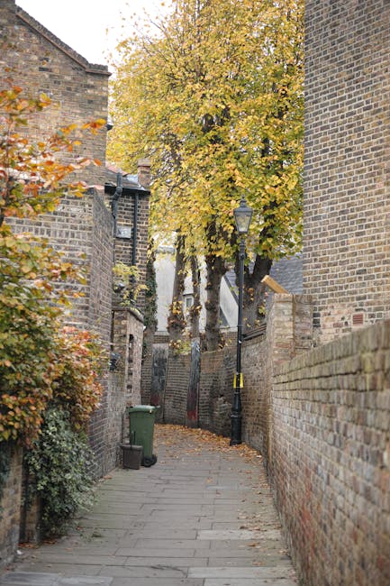 An alleyway with paved stone pathway surrounded by brick walls and buildings, featuring a large deciduous tree with yellow leaves at the end of the path. A black lamppost is visible along the side of the alley, and a green wheelie bin is positioned against a brick wall on the left. The scene depicts an outdoor urban environment typical of West Hampstead, where residential houses and narrow lanes are common, providing a backdrop for house removals and furniture transport activities. This setting may be part of a home relocation process managed by Man and Van West Hampstead, highlighting the typical environment for packing, loading, and transporting household items in a secure and organized manner.