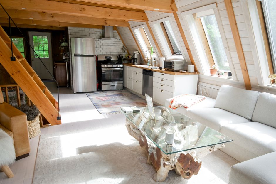 Interior view of a domestic loft space converted into a living area, featuring a white fabric sofa positioned against a wall with angled wooden beams and multiple skylight windows allowing natural light to fill the room. A rustic glass coffee table with a natural wood base is placed in front of the sofa, holding picture frames and decorative objects. To the left, part of a wooden staircase with black metal railing leads downstairs, with a woven storage basket underneath. The kitchen area in the background includes a stainless steel refrigerator, a stove with an extractor hood, white cabinetry with wooden countertops, and various small appliances like a microwave and a kettle on the work surface. The room has light-colored flooring and a neutral, modern decor style. The setting is within a house during a home relocation or packing process, typical of a project overseen by Man and Van West Hampstead, with objects neatly arranged and prepared for moving.