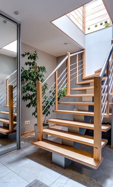 A wooden staircase with metal handrails inside a modern residential property, leading upwards to an open loft or upper level. The stairs are located near a large window or skylight, allowing natural light to illuminate the area. Adjacent to the stairs is a potted green plant, and a reflective glass door or mirror is visible on the left. The surrounding walls are painted in light neutral tones, and the floor is tiled. This interior scene depicts a typical home environment where furniture and objects might need to be carefully moved during a house relocation, with the staircase presenting challenges in moving large or bulky items. Man and Van West Hampstead, a moving specialist, might assist with such logistical issues during packing and furniture transport in NW6, West Hampstead.