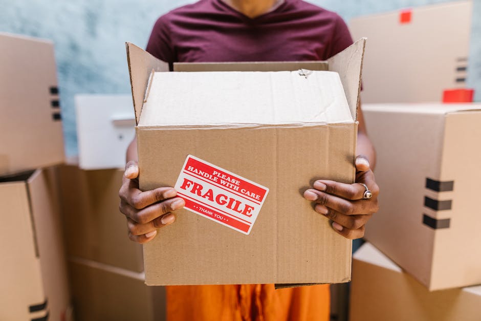 A person holding an open cardboard box marked with a red and white 'Fragile - Handle with Care' sticker, with their hands visible and wearing a silver ring on one finger. The individual is positioned inside a room surrounded by several other closed cardboard boxes, some with black tape securing the flaps. The background includes a partially visible white appliance or piece of furniture, and there is natural light illuminating the scene. This image depicts a home relocation process involving careful packing and handling of items, with the person preparing to load or transport the fragile box, which is part of the furniture transport and packing activities managed by Man and Van West Hampstead. The setting indicates a moving environment focused on house removals in the Westminster area, highlighting the importance of proper packing for efficient moving services.