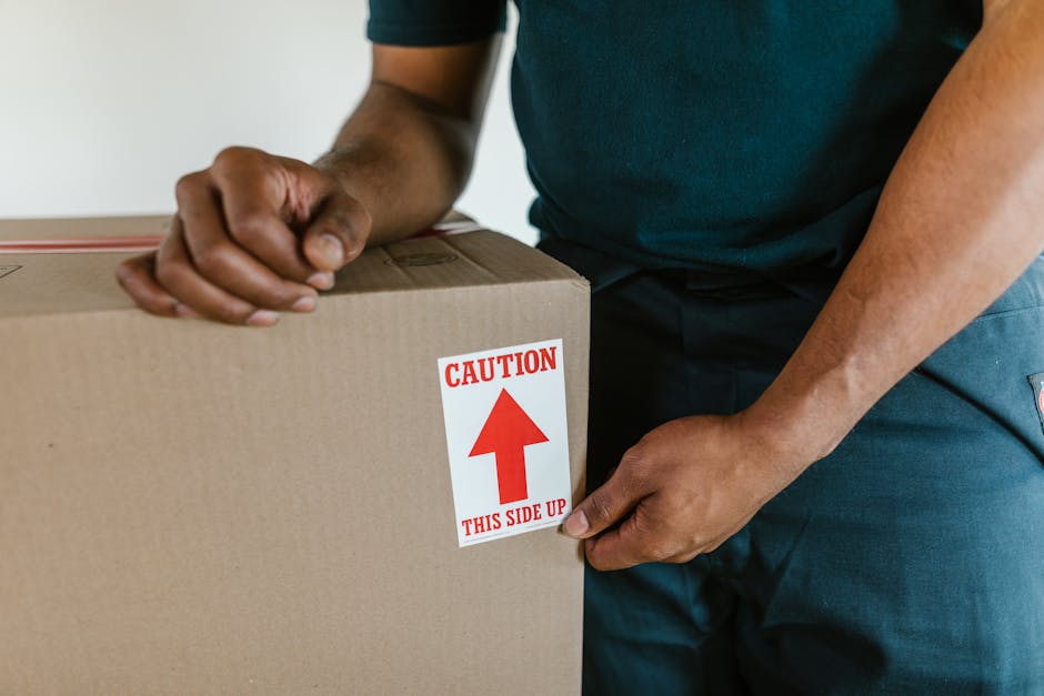 Close-up of a person wearing a dark green shirt and dark trousers handling a large cardboard box during a home relocation, inside a property near a doorway. The individual is using both hands to grip the box, which has a white and red label reading 'CAUTION THIS SIDE UP' with an upward arrow, indicating careful handling during the furniture transport process. The box appears to be sealed with packing tape, and in the background, part of the interior wall and floor are visible. The scene reflects the careful packing and loading activities involved in house removals, as performed by Man and Van West Hampstead, especially when dealing with delicate or heavy items requiring proper orientation and safety precautions during a house move.