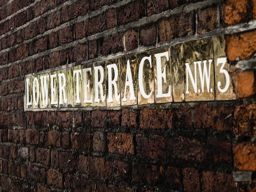 A close-up photograph of a wall-mounted street sign reading 'Lower Terrace N.W.3' on a weathered brick wall. The sign consists of multiple rectangular ceramic tiles with white lettering, showing signs of age such as dirt and minor discoloration. The bricks surrounding the sign are dark red and brown, with some mortar visible between them. The photograph captures the sign and wall at an angle, highlighting the texture of the bricks and the contrast between the sign's bright surface and the aged wall. This image depicts an urban residential area potentially in West Hampstead, illustrating typical signage seen during home relocations or furniture transport as part of house removal services by Man and Van West Hampstead, supporting the thematic focus on moving logistics and packing preparation for NW6 renters.