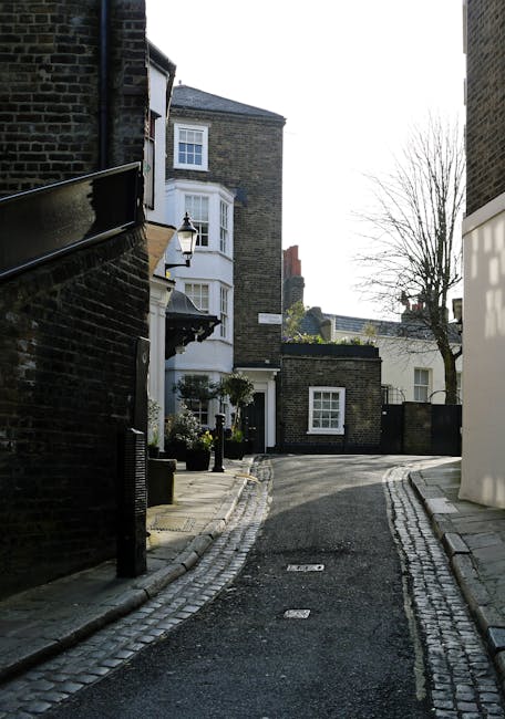 A narrow residential street featuring cobblestone paving and a slight curve, bordered by brick and white-painted buildings typical of West Hampstead. On the left, a black brick building with a small outdoor lantern next to a black fence. Potted plants with greenery are positioned near a doorway. On the right, a white building with a modern design. In the foreground, the street is clear, but further down, a large white van is partially visible, parked adjacent to a white building, with a truck load of packed cardboard boxes, furniture wrapped in plastic, and removals blankets visible in the open cargo area. This scene captures elements of home relocation and furniture transport, consistent with a professional house removals process. The environment is lit by natural daylight, and there are no visible people in the image. Man and Van West Hampstead services typically handle such packing, loading, and transportation activities related to student or household moves.