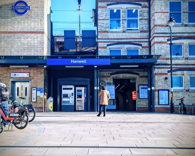 A woman dressed in a beige coat and black trousers stands outside the entrance of Hanwell station, which features a black canopy with the station name in white lettering on a blue background. The stat