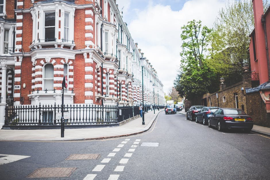 A quiet residential street in West Hampstead with a row of Victorian-style terraced houses featuring red brick facades, decorative white trim, and bay windows. The pavement runs along the front of these buildings, fenced by black metal railings with small decorative finials. Several parked cars, including sedans and hatchbacks, are lined up along the curb on the right side of the street. The street has white dashed road markings and is bordered by tall black bollards. In the background, a moving truck is visible, suggesting active home relocation or furniture transport. Green trees with fresh spring leaves shade parts of the street and partially obscure buildings further down the road. The sky is partly cloudy, with soft natural light illuminating the scene, conveying a typical day during a house removal or home packing and moving process. The area appears calm and orderly, with a focus on the urban architecture and street environment typical of West Hampstead, managed by Man and Van West Hampstead.